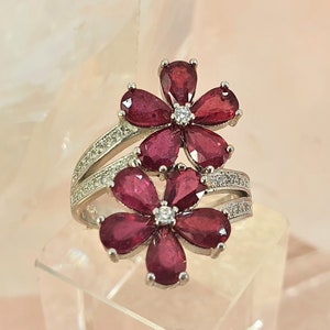 Puede incluir: Un anillo de plata con dos diseños de flores de rubí rojo. Las flores están hechas de rubíes en forma de pera y están rodeadas de diamantes blancos más pequeños.