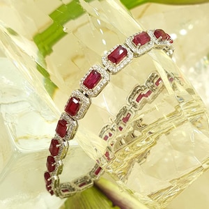 May include: A silver bracelet with a row of red square-shaped gemstones set in a silver frame. The bracelet is on a white surface with a green leaf.