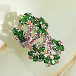 Peut inclure: Une bague en argent avec un motif floral composé de pierres précieuses vertes, roses et jaunes. La bague est posée sur une surface en marbre blanc.