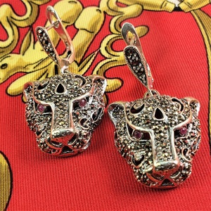 May include: A pair of silver dangle earrings featuring a detailed panther head design with pink gemstone eyes. The earrings are adorned with marcasite stones.