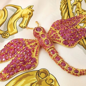 Puede incluir: Un broche de libélula de oro incrustado con rubíes rosas. Las alas y el cuerpo de la libélula están cubiertos de pequeños rubíes facetados.