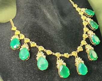 Op de afbeelding: Een gouden ketting met een ketting en meerdere groene edelstenen in druppelvorm. De ketting ligt op een zwarte fluwelen achtergrond.