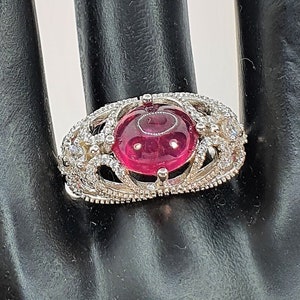 May include: A silver ring featuring a large, round, cabochon-cut, red gemstone surrounded by smaller, clear gemstones. The ring is displayed on a black, mannequin hand.
