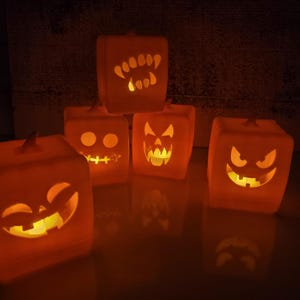 May include: A collection of glowing orange Halloween-themed cube-shaped lanterns. Each lantern features a different carved face, including fangs, a stitched mouth, and a sinister grin. The lanterns are lit from within, creating a warm, inviting glow.