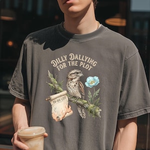 Puede incluir: Camiseta gris oscuro con una ilustración detallada de un pájaro posado en una rama, una flor azul y un pergamino con la inscripción "TO-DO LIST". El texto "Dilly Dallying for the Plot" está arqueado sobre el pájaro.