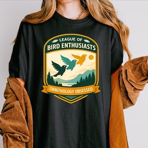 Puede incluir: Camiseta negra con un diseño gráfico colorido. El diseño incluye el texto "LEAGUE OF BIRD ENTHUSIASTS" y "ORNITHOLOGY OBSESSED". El diseño muestra tres pájaros volando sobre un paisaje con árboles y un sol. Prenda de vestir informal.