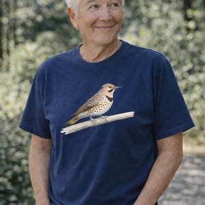 May include: Navy blue t-shirt featuring a detailed illustration of a Northern Flicker bird perched on a branch. The bird has a red nape, speckled breast, and yellow accents. The shirt is made of a soft, dark blue fabric.