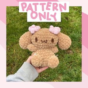 Puede incluir: Un conejito de crochet de color marrón, con lazos rosas en las orejas. El conejito tiene una cara sonriente con detalles bordados. El texto "PATTERN ONLY" se muestra en rosa encima del conejito.