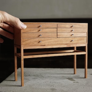 May include: A miniature wooden cabinet with six drawers and silver knobs. The cabinet is made of light brown wood and stands on four slender legs. A small shelf is located between the legs. The cabinet is a handcrafted item.