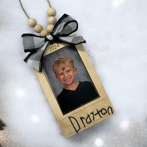 May include: A wooden ornament with a black and white checkered bow, a photo of a young boy, and the year 2024 written on it. The ornament is labeled "Drayton".