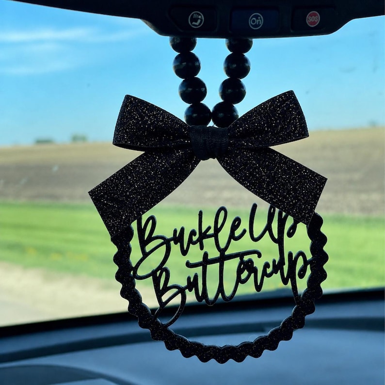 May include: Black car charm with a glittery bow and a circular frame. The frame features the words "Buckle Up Buttercup" in a cursive font. The charm hangs from a string of black beads, designed to hang from a car's rearview mirror.