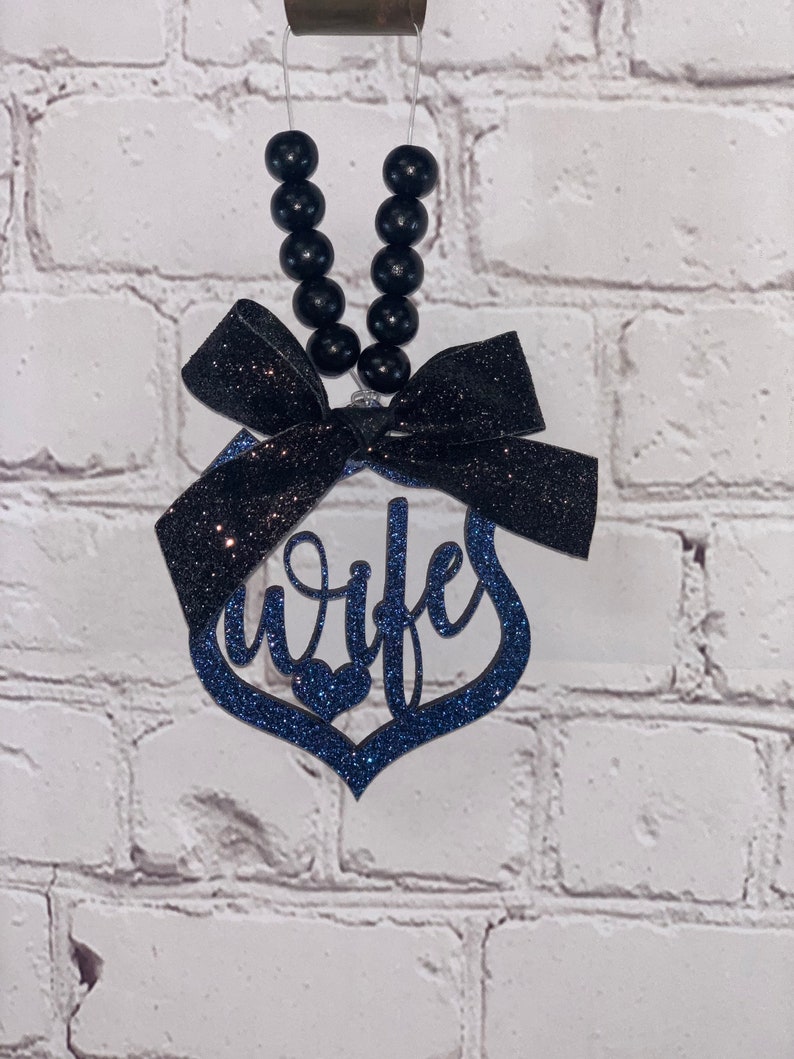 Pu&ograve; includere: Un ciondolo per auto in glitter blu e nero con la parola "moglie" in un design a forma di cuore. Il ciondolo &egrave; attaccato a una stringa di perline nere con un fiocco nero.
