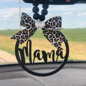 May include: A black glittery car charm with the word "Mama" in a cursive font, encircled by a black ring. It features a bow with a leopard print pattern and a white center, suspended by black beads.