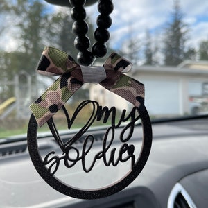 Peut inclure: Charme de voiture noir avec une finition paillet&eacute;e et un ruban camouflage. Le charme est un cercle avec les mots "J'aime mon soldat" &eacute;crits en cursive.