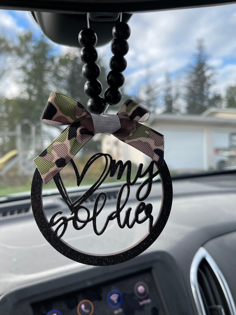 Peut inclure: Charme de voiture noir avec une finition paillet&eacute;e et un ruban camouflage. Le charme est un cercle avec les mots "J'aime mon soldat" &eacute;crits en cursive.