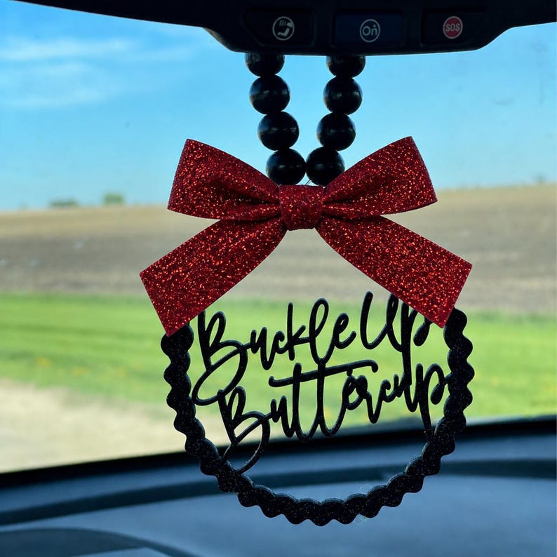 May include: A car charm with a red glitter bow and a black circular frame. The frame has the words "Buckle Up Buttercup" in a cursive font. The charm hangs from black beads.