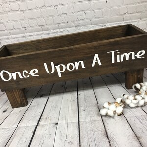 May include: A wooden box with the words "Once Upon A Time" painted in white on the front. The box is sitting on four short legs and is on a whitewashed wood floor.