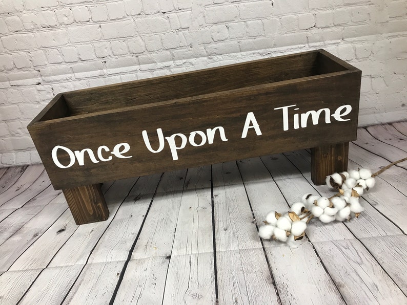 May include: A wooden box with the words "Once Upon A Time" painted in white on the front. The box is sitting on four short legs and is on a whitewashed wood floor.