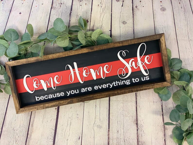 May include: A wooden sign with a black background and a red stripe across the middle. The sign reads "Come Home Safe because you are everything to us".