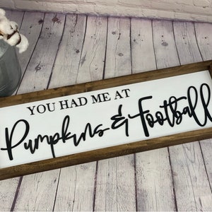 May include: A white wooden sign with a brown wood frame. The sign says "You had me at Pumpkins & Football" in black cursive lettering.