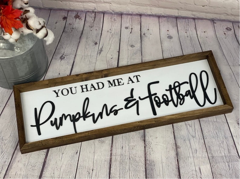 May include: A white wooden sign with a brown wood frame. The sign says "You had me at Pumpkins & Football" in black cursive lettering.