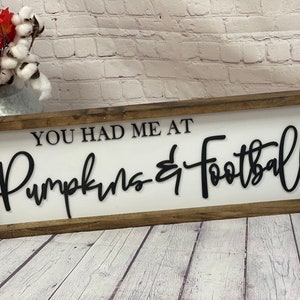 May include: A wooden sign with a white background and black lettering that reads "You had me at Pumpkins & Football".