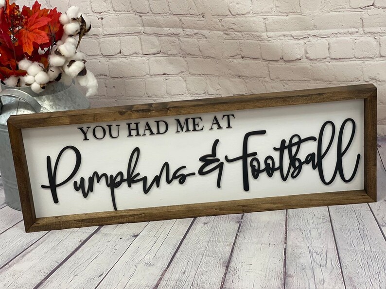 May include: A wooden sign with a white background and black lettering that reads "You had me at Pumpkins & Football".