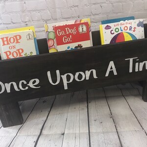 May include: A black wooden book holder with four legs, painted with white text that reads "Once Upon A Time". The holder is filled with four children's books: "Hop on Pop", "Go, Dog, Go!", "Lift-the-Flap Colors", and another book with a yellow cover.