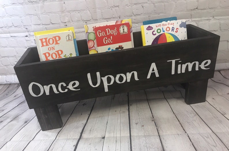 May include: A black wooden book holder with four legs, painted with white text that reads "Once Upon A Time". The holder is filled with four children's books: "Hop on Pop", "Go, Dog, Go!", "Lift-the-Flap Colors", and another book with a yellow cover.