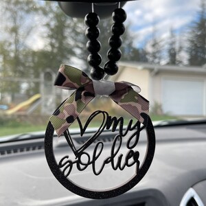 Peut inclure: Noeud en ruban camouflage noir et vert avec un cercle en bois noir paillet&eacute; et les mots "J'aime mon soldat" suspendu &agrave; un r&eacute;troviseur avec des perles noires.