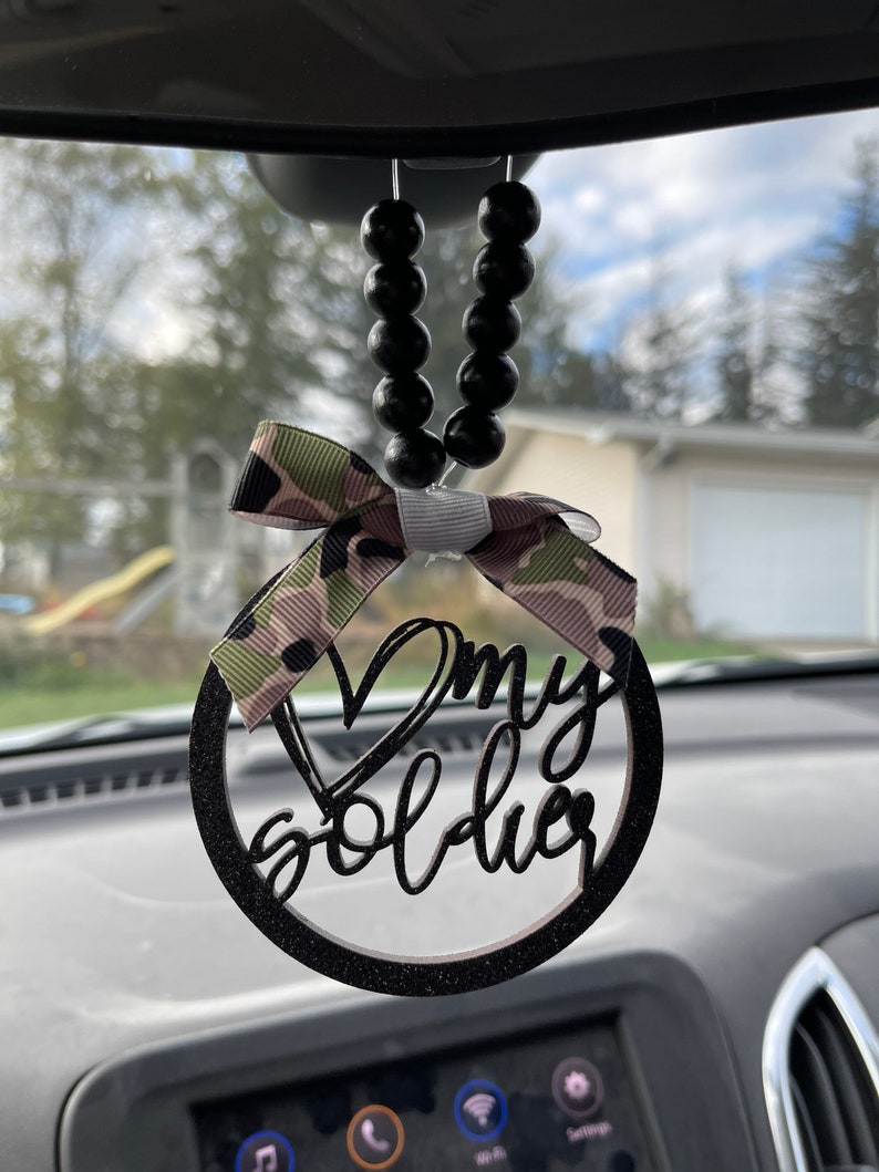 Peut inclure: Noeud en ruban camouflage noir et vert avec un cercle en bois noir paillet&eacute; et les mots "J'aime mon soldat" suspendu &agrave; un r&eacute;troviseur avec des perles noires.