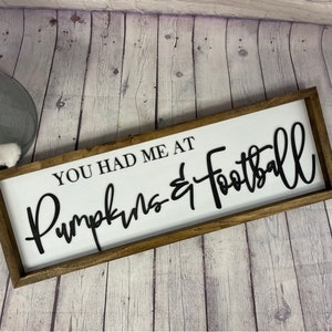 May include: A wooden sign with the text "You had me at Pumpkins & Football" in black lettering on a white background.