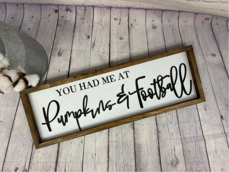 May include: A wooden sign with the text "You had me at Pumpkins & Football" in black lettering on a white background.