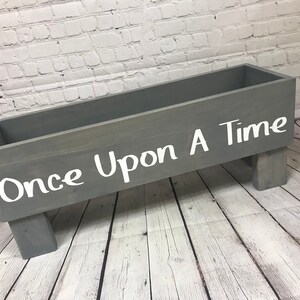 May include: A gray wooden box with the words "Once Upon A Time" written in white paint. The box is sitting on four short legs.