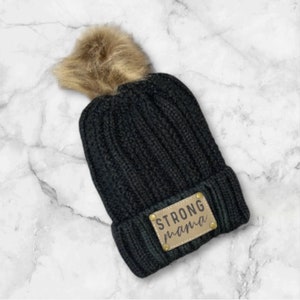 May include: Black knit beanie with a brown faux fur pom pom and a brown leather patch that says "STRONG mama".