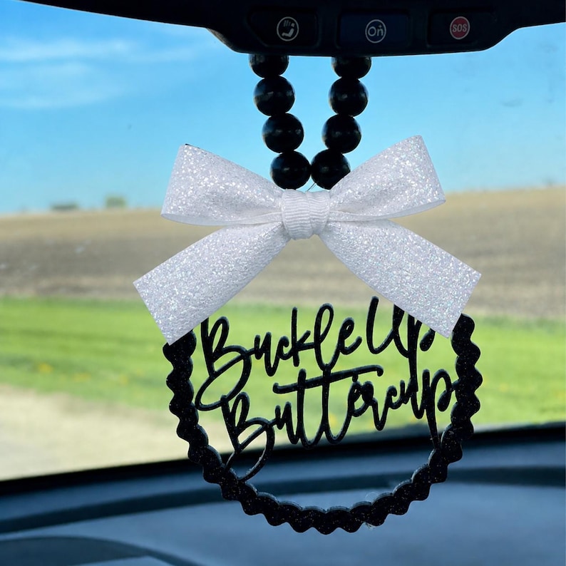 May include: A car charm with a white glitter bow and a black circular pendant. The pendant has the words "Buckle Up Buttercup" in a cursive font. The charm hangs from a string of black beads.