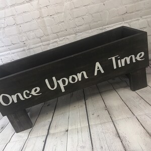 May include: A black wooden box with the words "Once Upon A Time" painted in white on the front. The box is sitting on a white wooden floor and has four legs.