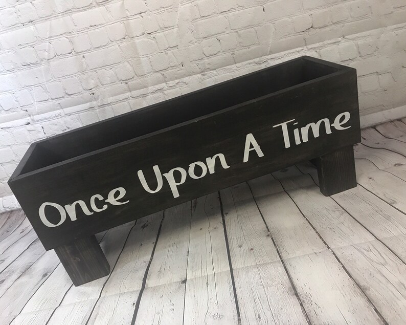 May include: A black wooden box with the words "Once Upon A Time" painted in white on the front. The box is sitting on a white wooden floor and has four legs.
