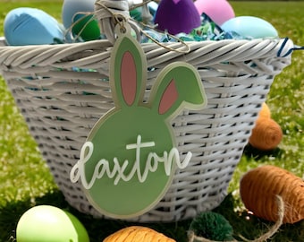 May include: A white wicker basket filled with colorful Easter eggs. A green bunny-shaped tag with the name "Laxton" is attached to the basket.