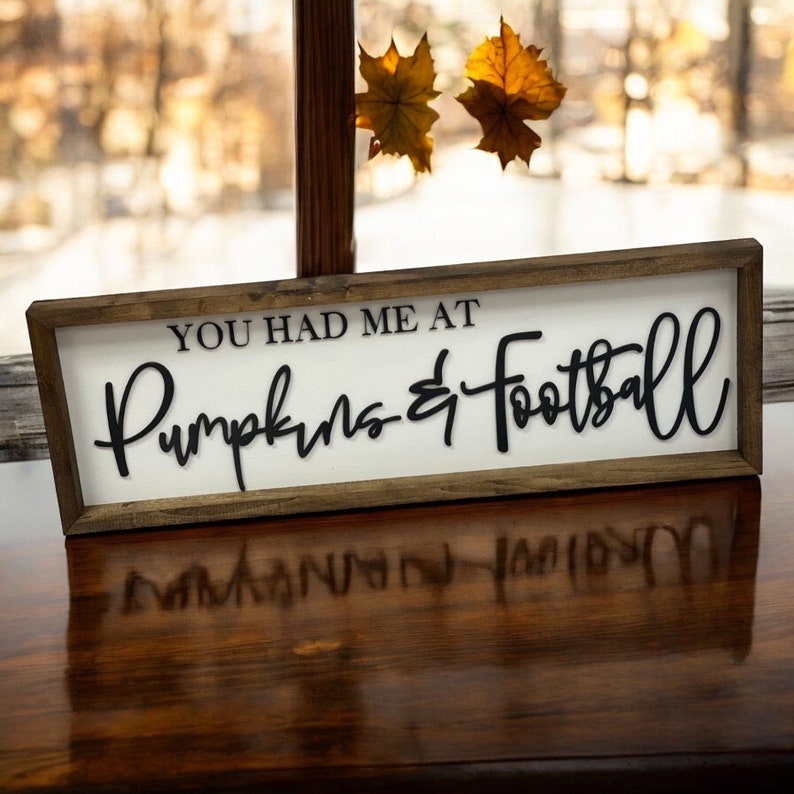 May include: A wooden sign with a white background and brown frame. The sign says "You had me at Pumpkins & Football" in black cursive lettering.