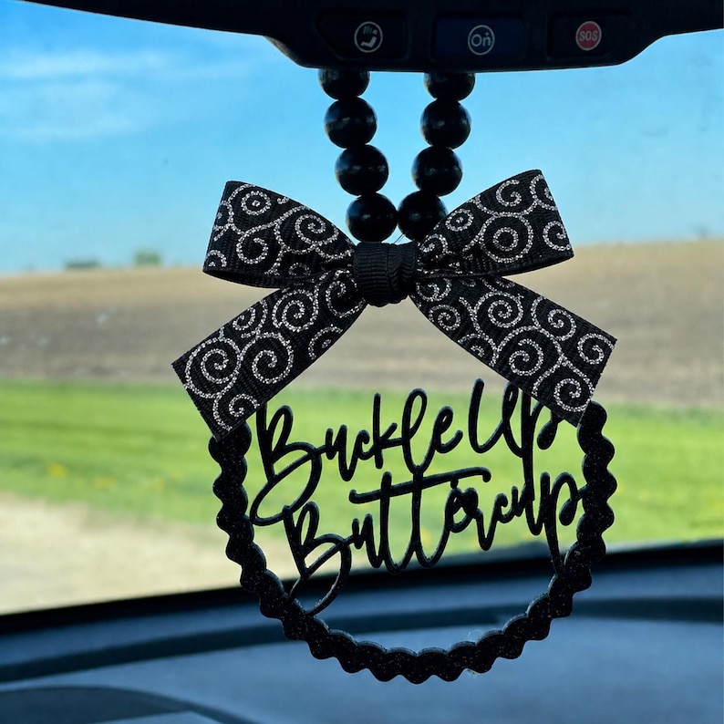 May include: Black car charm with a bow and the phrase "Buckle Up Buttercup". The bow has a black and silver swirl pattern. The charm hangs from a string of black beads.