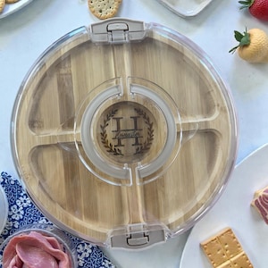 May include: A round bamboo serving tray with a clear plastic lid. The tray is divided into five sections and features the engraved name "Hamilton" with a decorative wreath. The tray is surrounded by crackers, cheese, and deli meats.