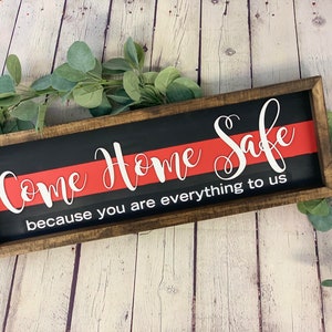 May include: A wooden sign with a black background and a red stripe across the middle. The sign reads "Come Home Safe because you are everything to us".