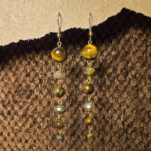 May include: A pair of dangle earrings featuring a combination of golden tiger eye beads and smaller, faceted beads in shades of green and brown. The beads are strung on gold-colored wire, with a simple hook closure. The earrings are displayed against a textured brown background.