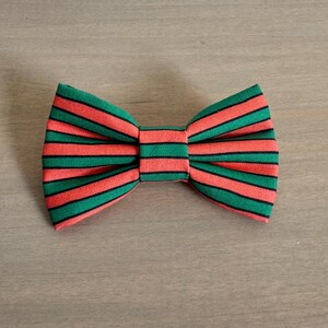 May include: A striped bow tie with alternating green and coral-red horizontal stripes. The bow tie is centered and appears to be made of fabric. The background is a light brown wooden surface.