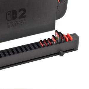 May include: A black game cartridge holder with a sliding mechanism, holding several game cartridges. The Nintendo Switch console is visible in the background. The holder has slots for the cartridges and red accents.