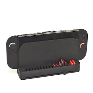 May include: Black Nintendo Switch docking station with a built-in game cartridge holder. The dock has a sleek design with a Switch console attached. The game cartridge holder features red and black accents. The Switch controllers have blue and orange accents.