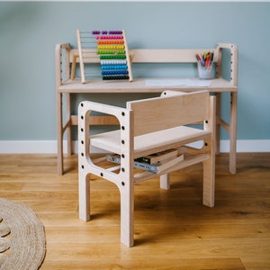 May include: A light-colored wooden desk and a matching wooden chair with black hardware. The desk holds a colorful abacus and a cup of pencils. The chair features a lower shelf holding books. The furniture has a smooth, natural wood finish.
