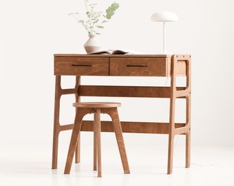 Könnte beinhalten: Ein heller Holzschreibtisch mit zwei Schubladen und einem Regal. Der Schreibtisch hat eine weiße Lampe und einen kleinen Holzhocker davor.