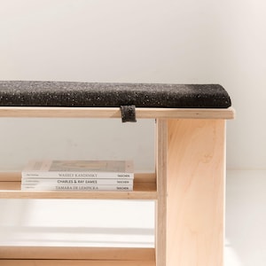 May include: A light-colored plywood bench featuring a dark gray, textured fabric cushion on top. The bench includes a lower shelf holding three books titled Wassily Kandinsky, Charles & Ray Eames, and Tamara de Lempicka, all published by Taschen.
